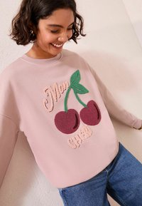 Felpa rosa chiaro con foglie verdi ricamate e due ciliegie burgundy; testo "Mon Chéri" in corsivo rosa sopra il design.