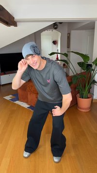 Szürke póló rózsaszín logóval, széles fekete nadrág, valamint kék, mintás sapka. A környezetben növény és modern berendezések láthatók.