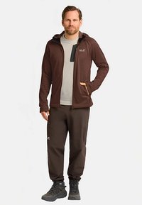 Veste marron à fermeture éclair avec capuche, chemise grise en dessous, pantalon marron assorti et chaussures de randonnée noires, dotées de poches et d'un design épuré.