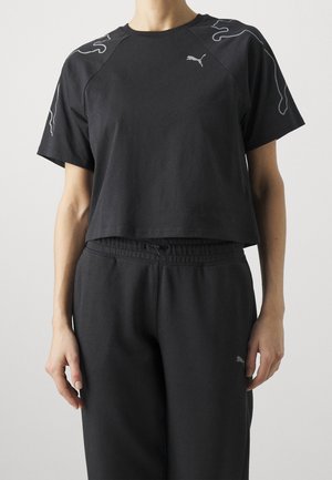 Frau trägt ein schwarzes Puma Cropped-T-Shirt mit abstrakten Linienmustern auf den Schultern und passende schwarze Jogginghosen.