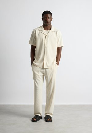 Homme debout contre un mur blanc portant une chemise crème à manches courtes boutonnée, un pantalon assorti, et des sandales noires avec les orteils ouverts et des brides croisées.