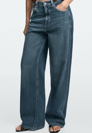 Personne portant un jean bleu foncé, taille haute, coupe ample, main dans la poche et sandales noires à bouts ouverts sur un fond uni.