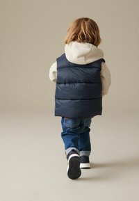 Gilet imbottito navy sopra una felpa grigia chiara, abbinato a jeans blu e sneakers nere. Il gilet ha cuciture orizzontali ed è leggermente imbottito.