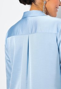 Camicia azzurro chiaro con una texture liscia, caratterizzata da una piega sul retro e un colletto classico. Orecchini argento visibili in primo piano.