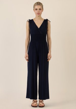 Frau mit blondem Haar, die einen ärmellosen marineblauen Jumpsuit mit geraffter Taille und Bindeträgern trägt, steht vor einem schlichten beigen Hintergrund.