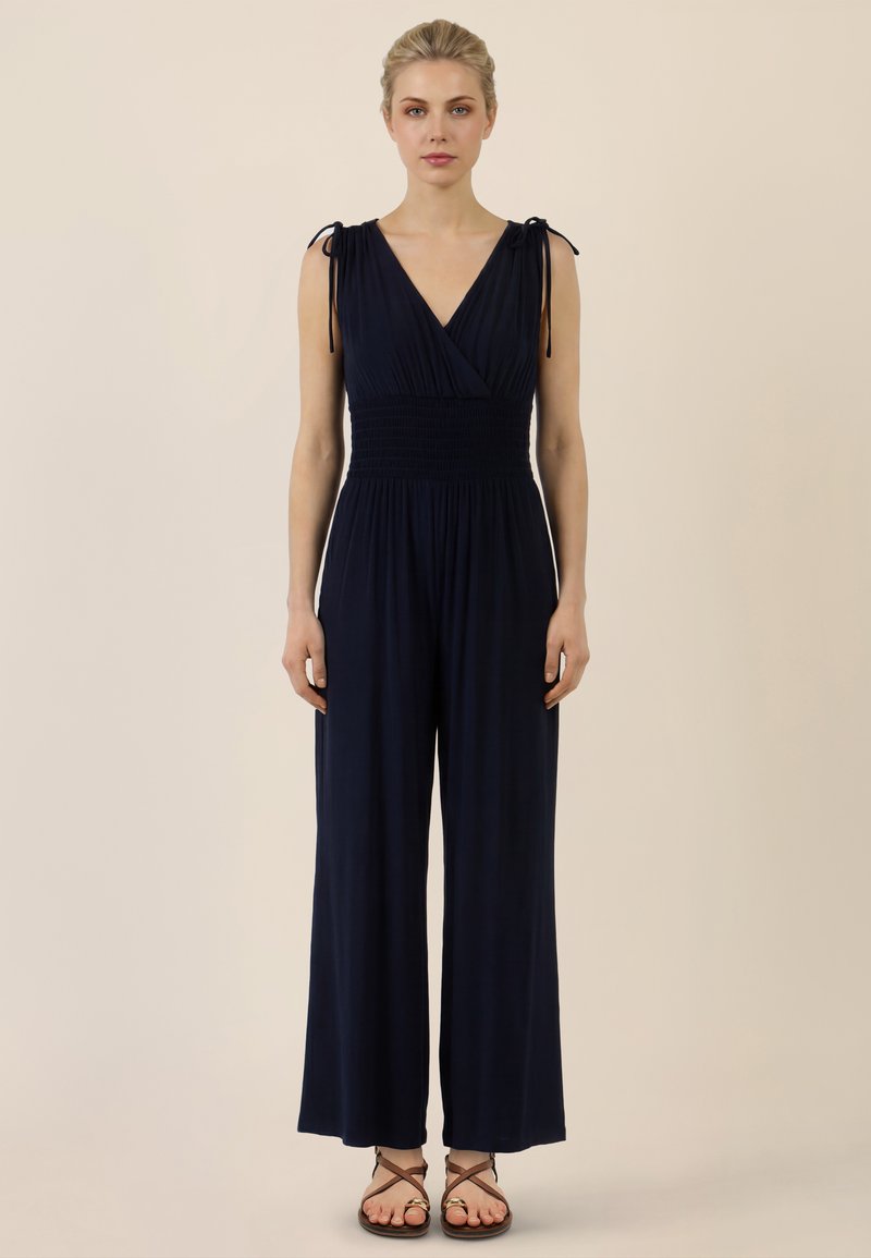 Kobieta o blond włosach ubrana w granatowy kombinezon bez rękawów ze ściąganym w talii paskiem i wiązanymi ramiączkami, stojąca na jednolitym beżowym tle.