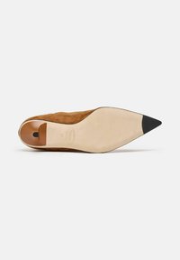 Zapato de ante marrón con punta afilada y suela beige lisa, y con una puntera negra. El interior cuenta con un corte redondeado para el talón. Talla 37.