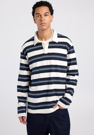 Jonge man met krullend haar, gekleed in een marineblauw-wit gestreept rugbyshirt met lange mouwen en donkere broek, staand tegen een effen achtergrond.