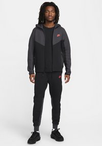 Schwarzer und grauer Nike Hoodie mit durchgehendem Reißverschluss, Taschen und Logo. Kombiniert mit passenden Jogginghosen mit Akzenten und schmal zulaufenden Säumen. Schwarze Schuhe.
