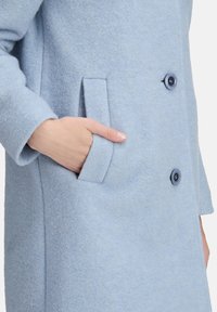 Cappotto in lana di colore azzurro chiaro con una finitura testurizzata, dotato di una tasca laterale e due bottoni scuri sul davanti. Design su misura, vestibilità classica.