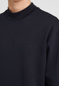 Marineblauer Pullover mit hohem Kragen, weich strukturiert und mit geprägtem "HUGO"-Logo auf der vorderen linken Seite.