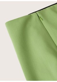Gros plan sur une jupe en tissu vert avec une fermeture éclair latérale dissimulée et une ceinture pliée sur fond blanc.