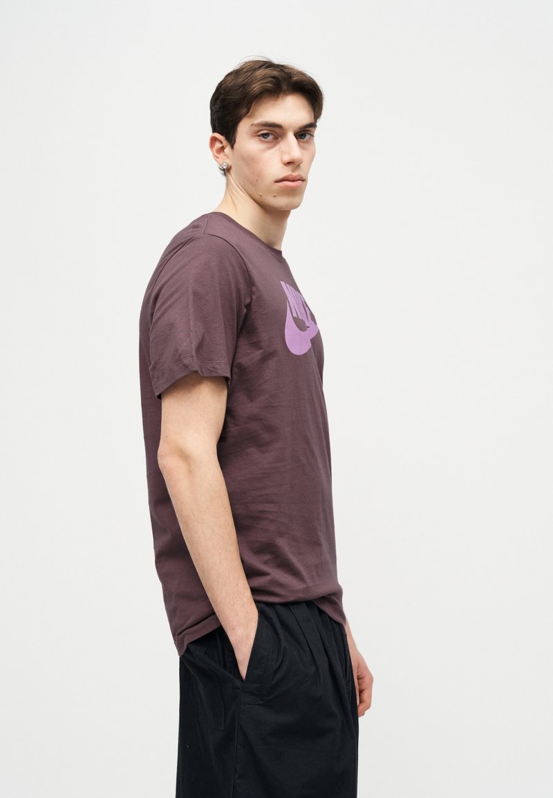 Jonge man met donker haar, gekleed in een paarse Nike T-shirt en zwarte korte broek, staand zijwaarts met handen in de zakken tegen een witte achtergrond.