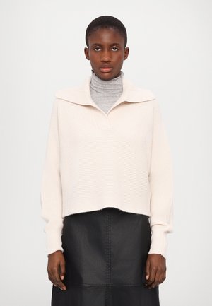 Pull en maille côtelée crème avec un large col, superposé à un col roulé gris. Jupe en cuir noir avec des coutures visibles. Design simple et structuré.