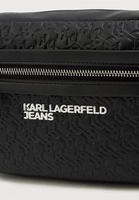 Saco de acessório preto com material texturizado apresentando um padrão de logótipos repetidos. Fecho de zíper prateado e nome da marca em branco exibido de forma proeminente.