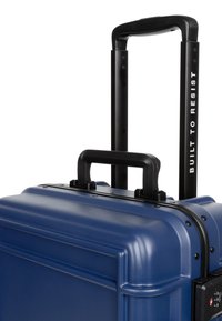 Kovakova valjačka torba u tamnoplavoj boji, s teleskopskom crnom ručkom, teksturiranom površinom i TSA odobrenom bravom sa strane.