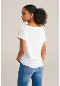 Balti tekstūruotas marškinėliai su žuvies kaulo raštu, suktomis rankovėmis, laisvo kirpimo, matomi iš nugaros. Suporuoti su mėlynomis džinsomis.