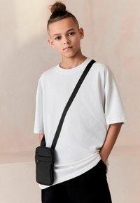 T-shirt blanc oversize à manches courtes, associé à un petit sac bandoulière noir avec fermeture éclair et sangle, sur un fond doux.