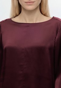 Femme aux longs cheveux blonds portant un chemisier bordeaux uni et brillant à encolure ronde, montrée des épaules à la poitrine sur fond clair.