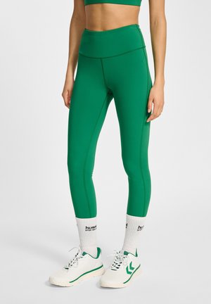 Gröna sportiga leggings i stretchmaterial med hög midja och sömdetaljer, i kombination med vita sportskor och strumpor.