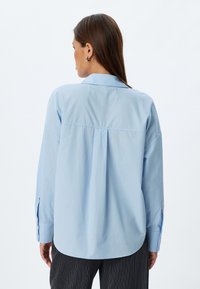 Camicia azzurro chiaro a maniche lunghe con colletto classico, caratterizzata da un dettaglio plissettato sul retro e polsini con bottoni, realizzata in un tessuto liscio.