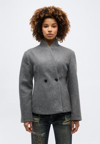 Γυναίκα με σγουρά μαλλιά φοράει γκρι διπλόχειρο μάλλινο σακάκι και φθαρμένα τζιν με διακοσμητικές τρουκς, στέκεται μπροστά σε λευκό φόντο.