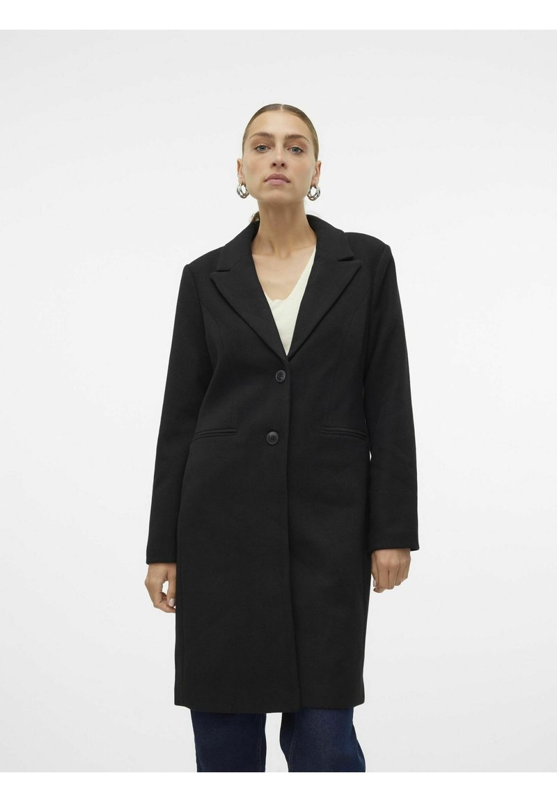 Abrigo largo negro con collar de solapa, dos botones y bolsillos laterales. Corte entallado y tejido texturizado; se lleva sobre una camisa ligera.