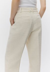 Gros plan sur une personne portant un pantalon texturé de couleur crème, mains dans les poches, associé à un haut blanc sans manches.