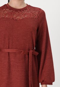 Rød strikket kjole med blonde detaljer på yoken, lange puffede ermer og belteknute i midjen. Strukturert stoff fullfører designet.