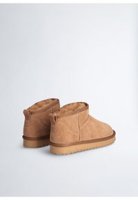 Stivaletti corti in suede color tan con una soffice foderatura in pelliccia e una suola in gomma. Presentano cuciture a vista e un piccolo logo sul lato.