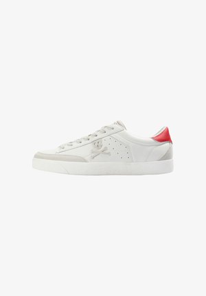 Zapatilla baja blanca con acentos de ante gris claro, una lengüeta trasera roja, logo de calavera y huesos cruzados en el lateral, y suela de goma blanca.