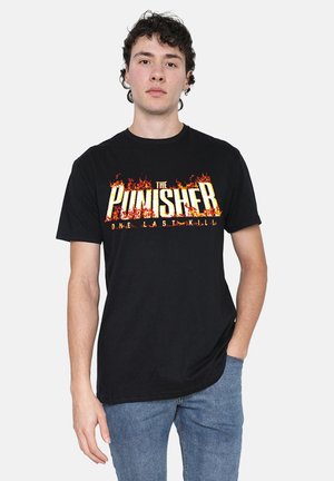 Junger Mann mit dunklen lockigen Haaren, der ein schwarzes The Punisher T-Shirt mit Flammen und blaue Jeans trägt, steht mit einer Hand in der Tasche.