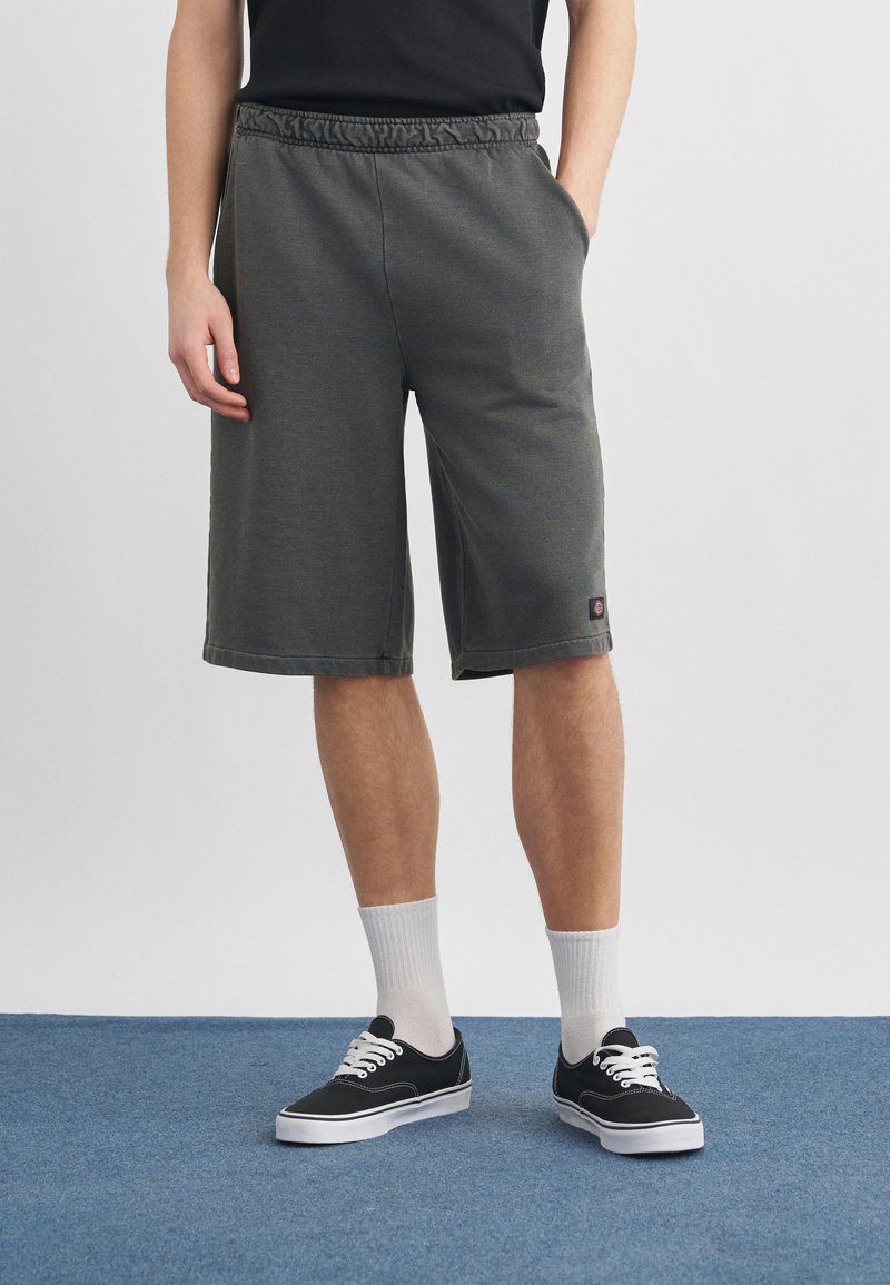 Persona usando pantalones cortos de color gris oscuro hasta la rodilla, zapatillas negras con cordones blancos, calcetines blancos y una camiseta negra, de pie sobre una alfombra azul.