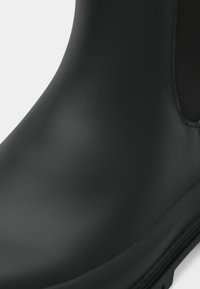 Botina de borracha preta com acabamento mate e suave, apresentando uma forma arredondada e painéis laterais elásticos para fácil calce. Sola texturizada para melhor aderência.