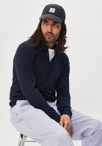 Marineblaues Langarm-Polo-Shirt mit Kragen und zwei Knöpfen, kombiniert mit hell gestreiften Hosen und einer schwarzen Kappe mit einem Logo.