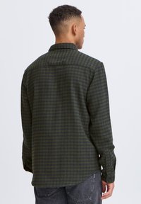 Mand iført mørkegrøn og marineblå ternet langærmet skjorte med sorte jeans, stående med ryggen mod en ensfarvet lys baggrund.