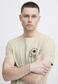 Lys beige t-skjorte med rund hals og korte ermer. Frontgrafikk med svart sol og tekst. Glatt bomullstekstur, casual passform.