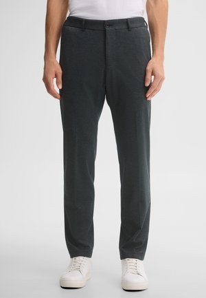 Uomo che indossa pantaloni dritti grigio scuro, sneakers bianche e camicia bianca infilata, su uno sfondo chiaro e uniforme.