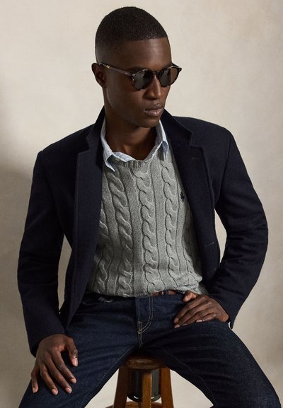 Uomo con occhiali da sole scuri, blazer blu navy, maglione grigio a trecce e jeans blu, seduto su uno sgabello di legno davanti a uno sfondo semplice.
