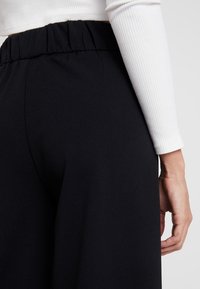 Czarne spodnie o szerokich nogawkach, z luźnym elastycznym pasem. Tkanina wydaje się gładka o miękkiej fakturze.