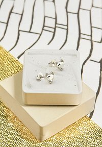 Söta kattformade silvriga studs visade på en matt betongfat, placerade på en gyllen texturerad yta med ett geometriskt mönster.
