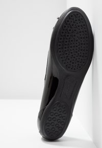 Chaussure noire brillante avec semelle en caoutchouc comportant des trous de ventilation ronds et une texture antidérapante. Les détails de couture sur les côtés renforcent le design.