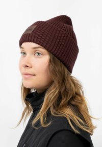Maroon knitted beanie with a ribbed texture and leather patch detail, worn by a person with wavy hair, paired with a black vest.