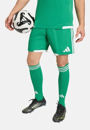 Mężczyzna w zielonym stroju piłkarskim trzyma czarno-białą piłkę Adidas, ubrany w pasujące skarpety i czarne korki, stoi na białym tle.