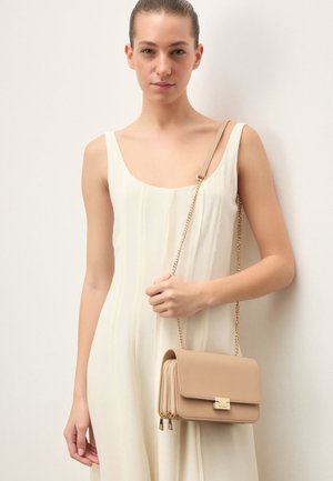 Žena v bielych šatách bez rukávov, ktorá drží béžovú kabelku crossbody so zlatým reťazovým remienkom na jednoduchom svetlom pozadí.