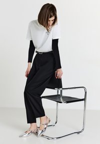 Pantalon noir taille haute, T-shirt blanc avec imprimé, chaussures argentées à bout pointu, chaise en chrome. Tissu texturé et lignes épurées.