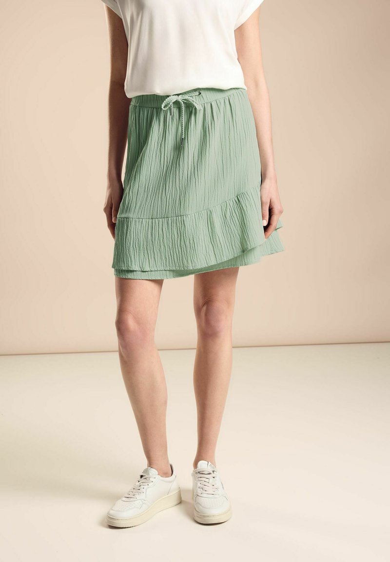 Gonna verde plissettata con vita regolabile, caratterizzata da un orlo stratificato. Indossata con sneaker bianche su uno sfondo neutro.