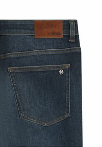 Jeans de mezclilla azul oscuro con una etiqueta de cuero marrón que presenta "BOSS" grabado. Incluye un bolsillo trasero con un detalle de logo bordado.