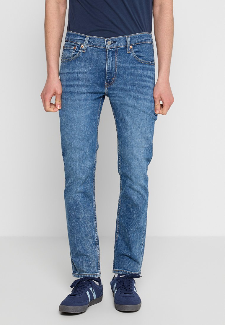 Jeans de mezclilla azul con un ajuste recto, ligera decoloración, cinco bolsillos y cierre de botón. Llevados con zapatillas navales.