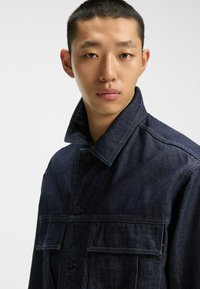 Mørk denimjakke med klassisk krave, forlommer og knaplukninger; udviser en robust stoftekstur og skræddersyet pasform.